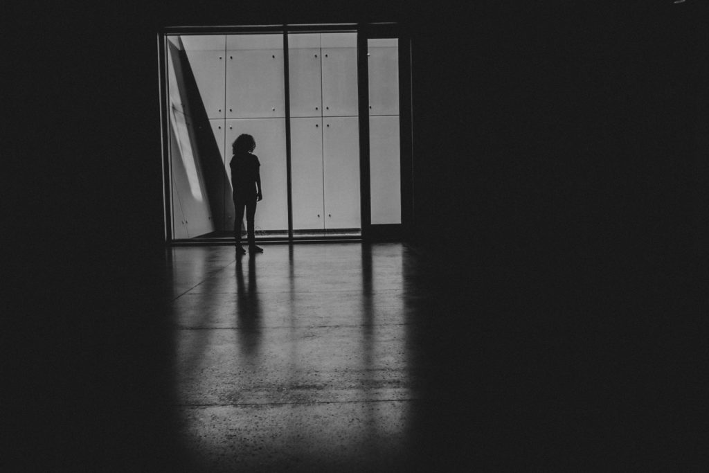 A black-and-white silhouette of a woman standing at the end of a hallway, facing away from the camera.